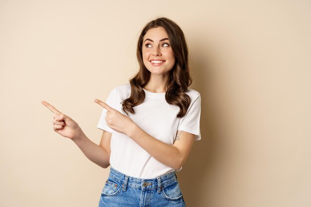 Elegante giovane donna caucasica sorridente che punta il dito a sinistra mostrando pubblicità offerta promo standi...