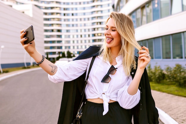 Elegante giovane donna che indossa un abito blu scuro alla moda, in posa vicino a edifici moderni, accessori alla moda, facendo selfie e mostrando la lingua, stato d'animo positivo.