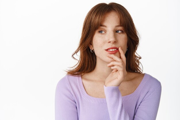 Elegante giovane donna con make up labbra rosse che guarda da parte sorridente premuroso e meditando in piedi su sfondo bianco in camicetta viola