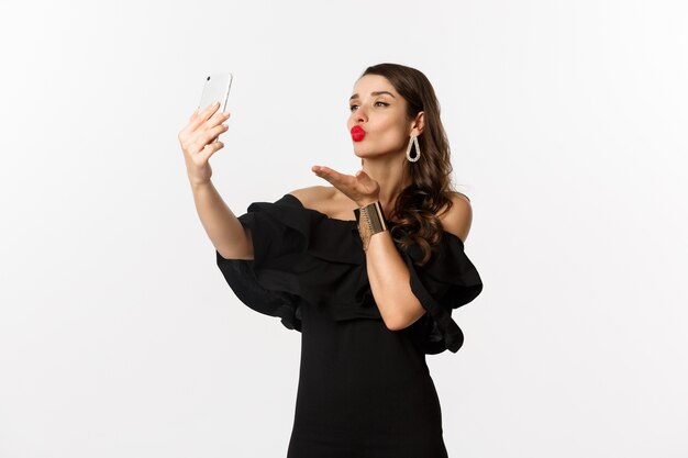 Elegante giovane donna in abito nero, festa e prendendo selfie sul cellulare, inviando un bacio d'aria alla fotocamera dello smartphone, in piedi su sfondo bianco.
