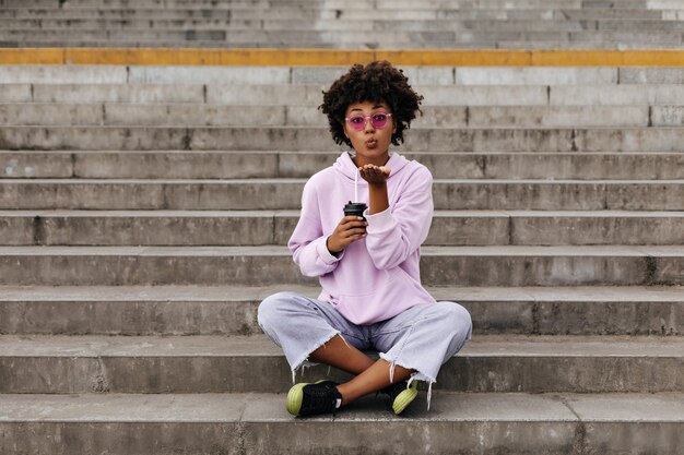 Elegante giovane donna in jeans, felpa con cappuccio rosa e occhiali da sole colorati si bacia, tiene in mano una tazza di caffè e si siede sulle scale all'aperto