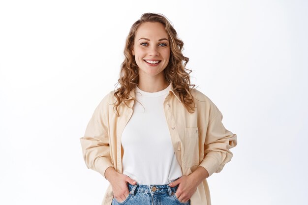 Elegante giovane donna moderna con una pettinatura bionda, che si tiene per mano nelle tasche dei jeans e sorride spensierata, sembra sicura e determinata, muro bianco