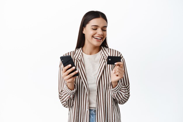 Elegante giovane donna moderna sorridente, guardando la carta di credito in plastica, pagando online con il telefono cellulare, facendo acquisti nell'app per smartphone, muro bianco