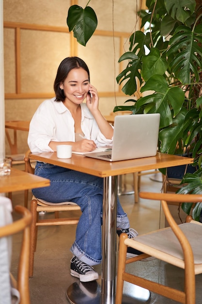 Elegante giovane donna risponde alla telefonata seduta al bar con il computer portatile che lavora e parla sullo smartphone