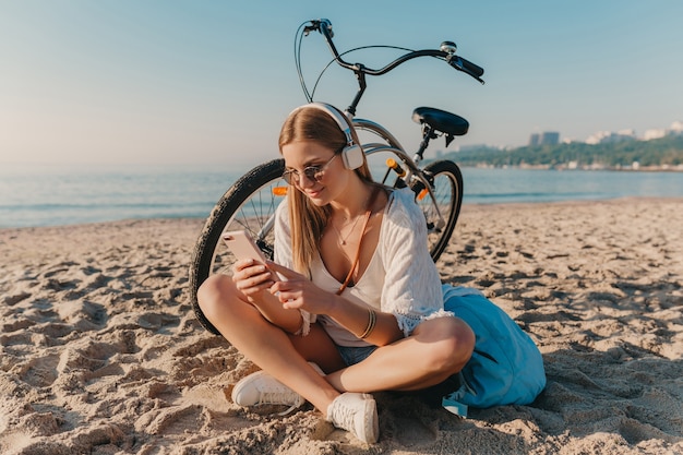 Elegante giovane donna sorridente bionda attraente che si siede sulla spiaggia con la bicicletta in cuffie che ascolta la musica