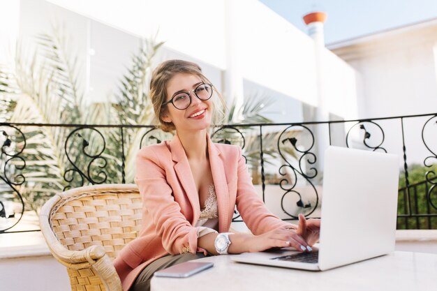 Elegante giovane donna sorridente e lavorando al computer portatile, ragazza allegra che digita sulle tastiere, seduto sulla sedia di vimini in un accogliente caffè all'aperto. Indossare occhiali eleganti, giacca rosa, camicetta beige, orologi bianchi.