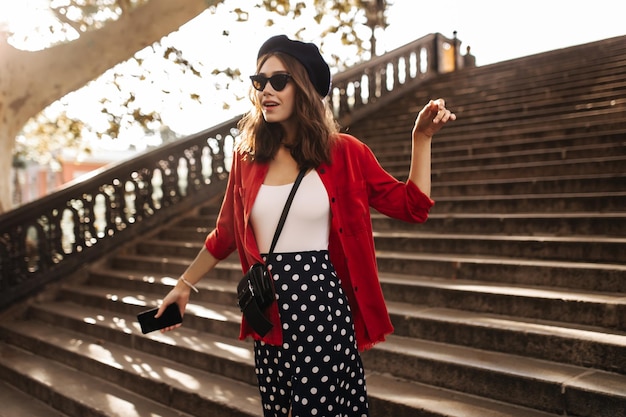 Elegante giovane Parisienne con capelli ondulati bruna, occhiali da sole neri, gonna bianca a pois e camicia rossa in piedi sulle scale della città e guardando dritto Sfondo autunnale caldo all'inizio