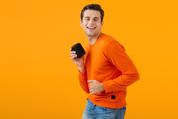 Elegante giovane sorridente con un maglione arancione che tiene in mano un altoparlante wireless felice ascolto di musica divertendosi