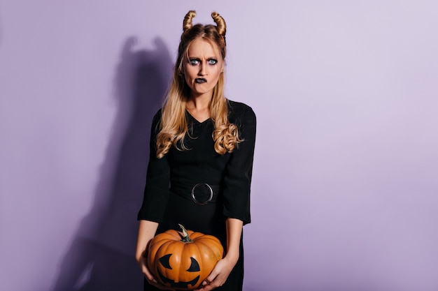 Elegante giovane strega che tiene la zucca di Halloween. Ragazza vampiro triste con trucco scuro in posa sulla parete viola.