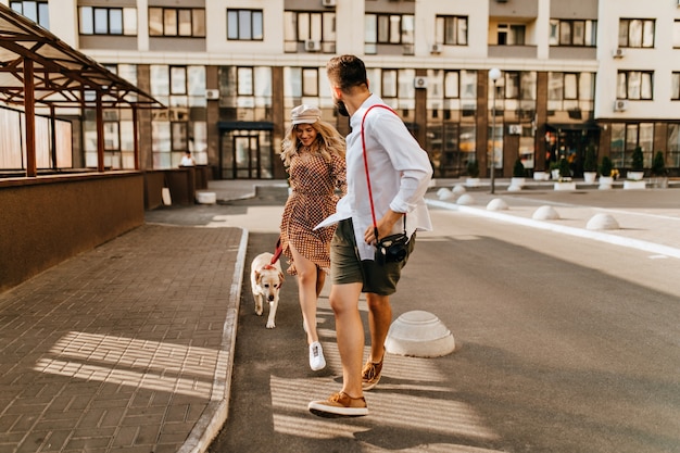 Elegante marito e moglie in abiti estivi corrono e giocano con il loro cane sullo sfondo del condominio. L'uomo in camicia leggera tiene la sua amata mano e trasporta la fotocamera.