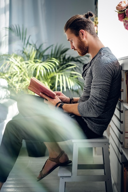 Elegante maschio barbuto che legge un libro in una stanza con piante verdi.