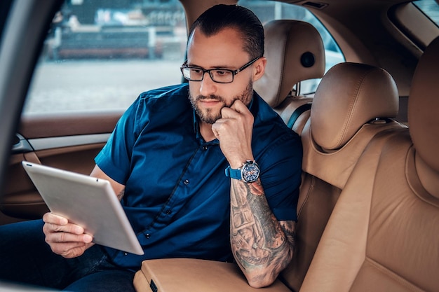 Elegante maschio barbuto in occhiali con tatuaggio sul braccio utilizzando un tablet PC portatile sul sedile posteriore di un'auto.