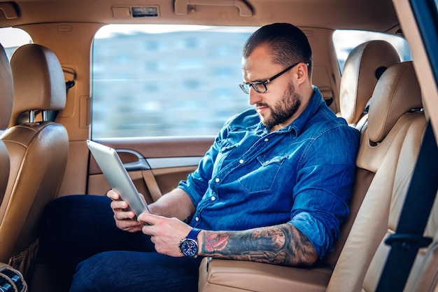 Elegante maschio barbuto in occhiali con tatuaggio sul braccio utilizzando un tablet PC portatile sul sedile posteriore di un'auto.