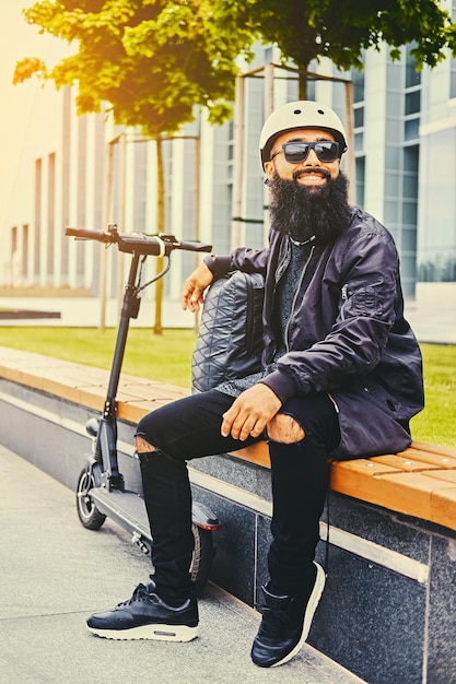 Elegante maschio barbuto in occhiali da sole che si rilassa su una panchina dopo aver guidato in scooter elettrico in centro.