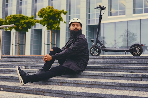 Elegante maschio barbuto su un gradino sopra lo sfondo di uno scooter elettrico.