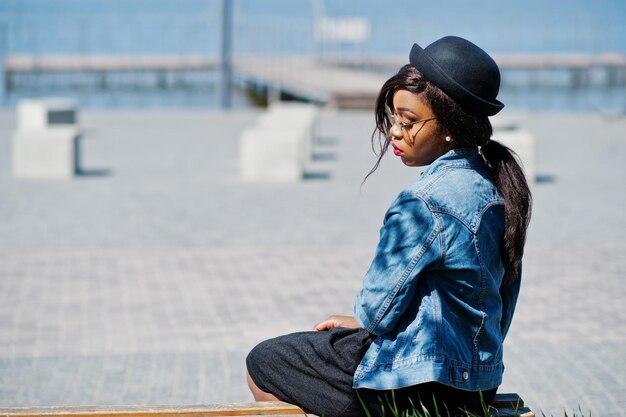 Elegante modello afroamericano in giacca di jeans cappello occhiali e gonna nera poste all'aperto