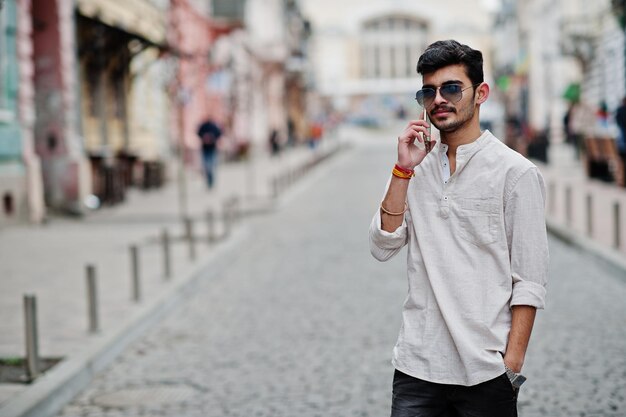 Elegante modello indiano uomo in abiti casual e occhiali da sole posato all'aperto in strada dell'India e parlando al telefono cellulare