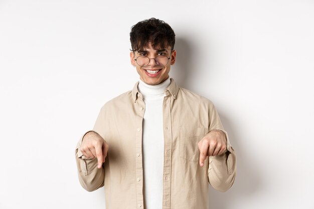 Elegante modello maschile hipster con gli occhiali che puntano le dita verso il basso, sorridendo soddisfatto alla telecamera, consigliando il prodotto, in piedi su sfondo bianco.