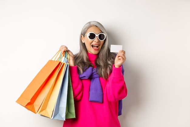 Elegante nonna asiatica in occhiali da sole che va a fare shopping durante i saldi delle vacanze, con in mano sacchetti di carta e carta di credito in plastica, in piedi su sfondo bianco.
