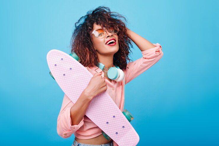 Foto gratuito elegante ragazza africana in camicia rosa che indossa grandi cuffie e occhiali da sole alla moda, sorridente. ritratto dell'interno della giovane signora nera con l'acconciatura riccia in posa con lo skateboard.