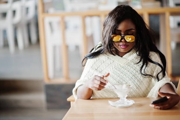 Elegante ragazza afroamericana in occhiali da sole posata al caffè moderno e mangiare il gelato