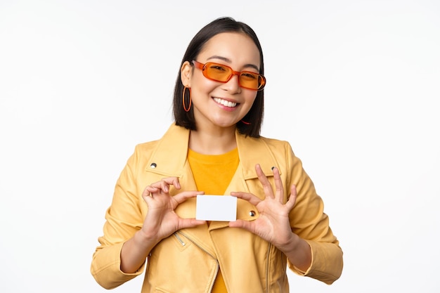 Elegante ragazza asiatica attraente in occhiali da sole mostrando carta di credito e sorridente in piedi felice su sfondo bianco per studio Copia spazio