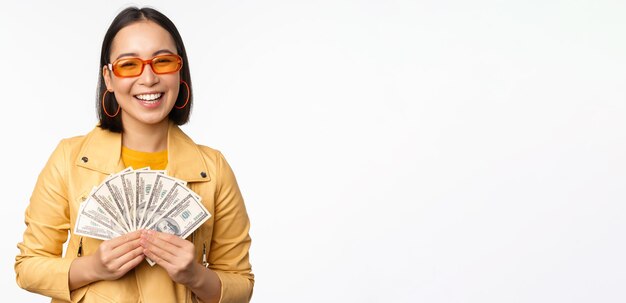 Elegante ragazza asiatica sorridente in possesso di denaro contante mostrando dollari e celebrando in piedi su sfondo bianco Copia spazio