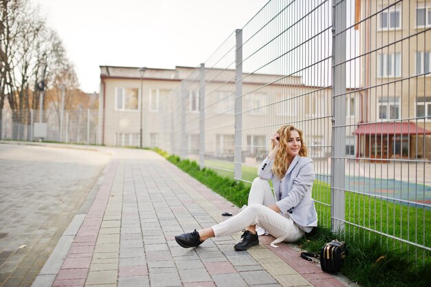 Elegante ragazza bionda riccia indossa su bianco in posa contro il recinto