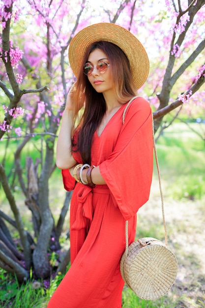 Elegante ragazza bohémien in posa in abito di corallo e cappello di paglia vicino al ciliegio in fiore nel parco di primavera.