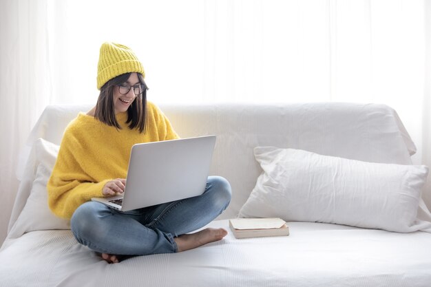 Elegante ragazza bruna allegra in occhiali e un cappello si siede sul divano a casa e lavora a un computer portatile