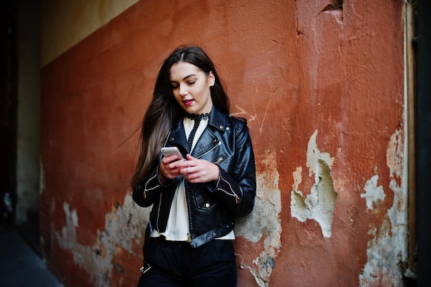 Elegante ragazza bruna indossare su giacca di pelle e pantaloncini con il cellulare a portata di mano contro il muro arancione