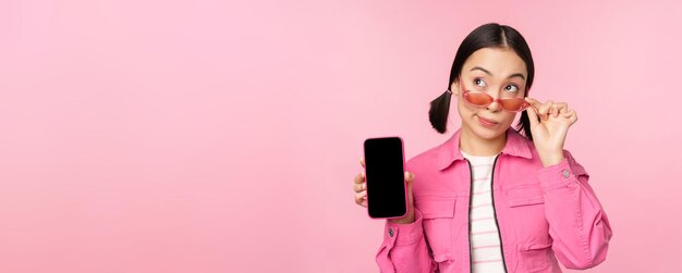 Elegante ragazza coreana giovane donna in occhiali da sole che mostra l'interfaccia dell'app del telefono cellulare dello schermo dello smartphone o il sito Web in piedi su sfondo rosa