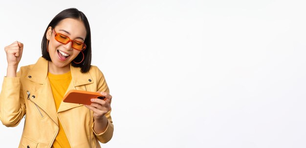 Elegante ragazza coreana in occhiali da sole che gioca al videogioco mobile ridendo e sorridendo mentre si utilizza lo smartphone in piedi su sfondo bianco