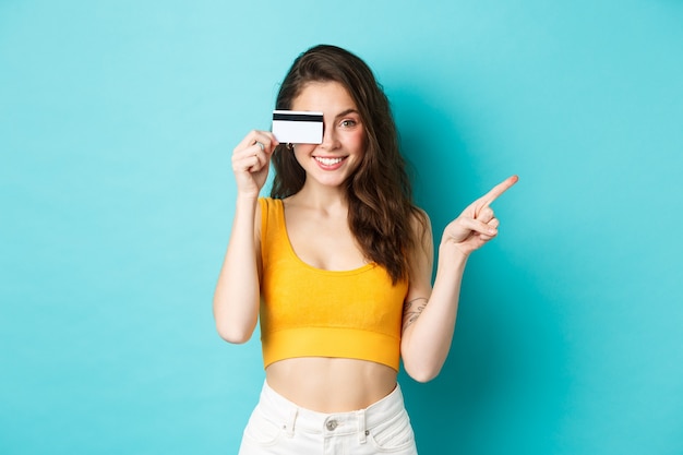 Elegante ragazza estiva con carta di credito rivolta da parte, mostrando il tuo logo sullo spazio della copia sul lato destro, in piedi su sfondo blu