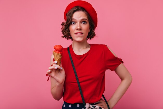 Elegante ragazza francese con il manicure bianco che tiene il gelato. Tiro al coperto di emotiva signora dai capelli scuri in vestiti rossi che mangia il suo dessert.