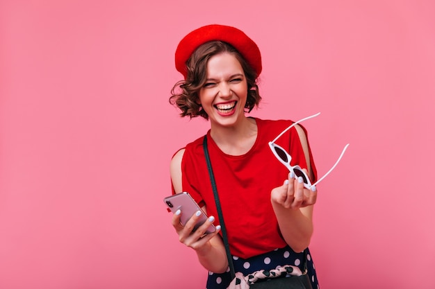 Elegante ragazza francese con occhiali da sole in mano e sorridente. Felice donna dai capelli scuri in abito rosso che ride.