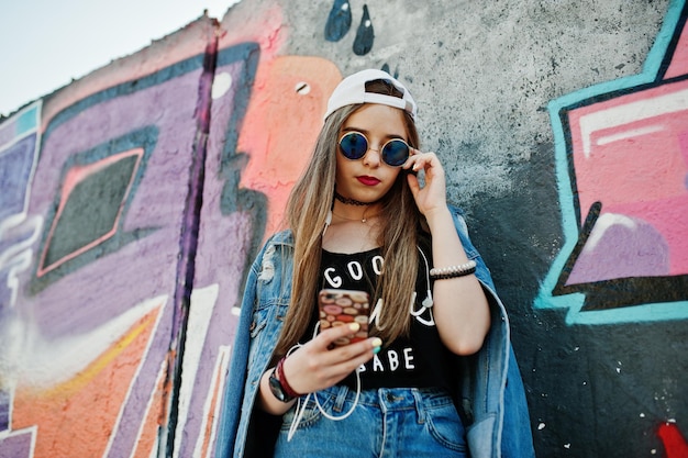 Elegante ragazza hipster casual con occhiali da sole e jeans con cappuccio indossa musica d'ascolto dalle cuffie del telefono cellulare contro il grande muro di graffiti