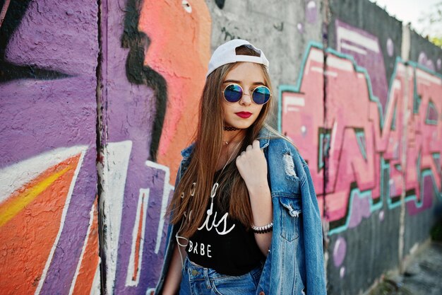 Elegante ragazza hipster casual con occhiali da sole e jeans con cappuccio indossa musica d'ascolto dalle cuffie del telefono cellulare contro il grande muro di graffiti