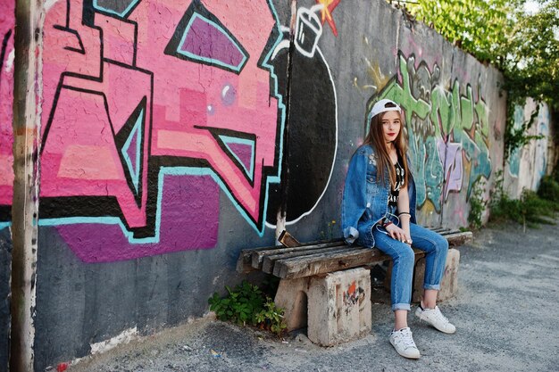 Elegante ragazza hipster casual in berretto e jeans indossa musica d'ascolto dalle cuffie del telefono cellulare contro il grande muro di graffiti con la bomba