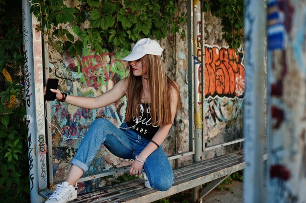 Elegante ragazza hipster casual in berretto e jeans indossa musica d'ascolto dalle cuffie del telefono cellulare contro il grande muro di graffiti