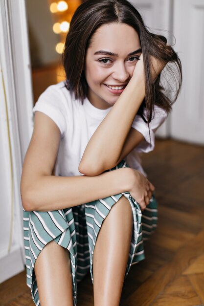 Elegante ragazza in posa in pigiama nel buon fine settimana mattina. Bruna donna caucasica seduta sul pavimento con un bel sorriso.