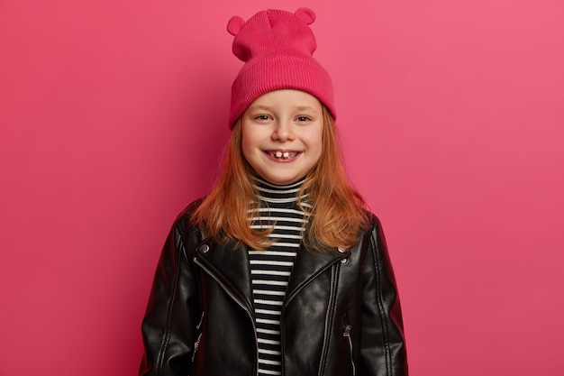Elegante ragazza rossa positiva sorride ampiamente, ha i denti mancanti, indossa una giacca di pelle alla moda e un cappello rosa, si gode una bella giornata, trascorre il fine settimana con i genitori, è di ottimo umore, posa al coperto