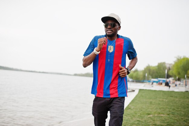 Elegante ragazzo afroamericano che corre contro il lago indossa una maglietta da calcio e occhiali da sole Ritratto di uomo sportivo nero