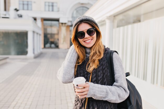 Elegante ritratto gioiosa donna alla moda con lunghi capelli biondi, cappello lavorato a maglia, caldo maglione di lana e occhiali da sole moderni che camminano nel centro della città. Viaggiare con il caffè per andare, felicità. Posto per il testo.