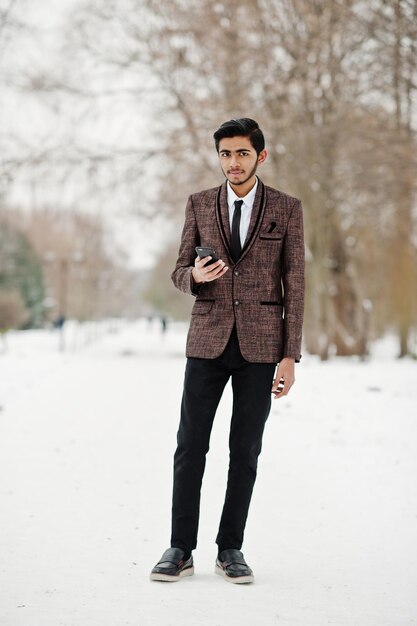 Elegante studente indiano in abito marrone e occhiali posato in una giornata invernale all'aperto con il telefono cellulare a portata di mano