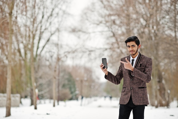 Elegante studente indiano in abito marrone e occhiali posato in una giornata invernale all'aperto con il telefono cellulare a portata di mano