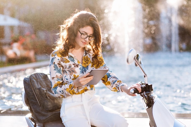 Elegante studentessa in occhiali alla moda che legge attività agghiacciante accanto alla fontana della città dopo le lezioni. Ritratto all'aperto di una romantica ragazza riccia con tablet e zaino in pelle nera che fa i compiti.