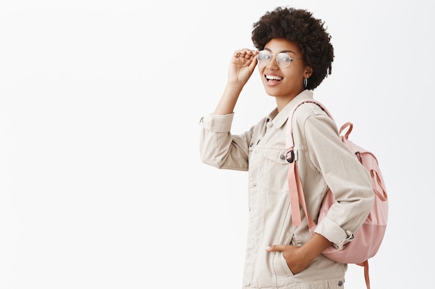 Elegante turista femminile di bell'aspetto con pelle scura e acconciatura afro che controlla gli occhiali sugli occhi e si gira mentre si trova di profilo sopra il muro grigio