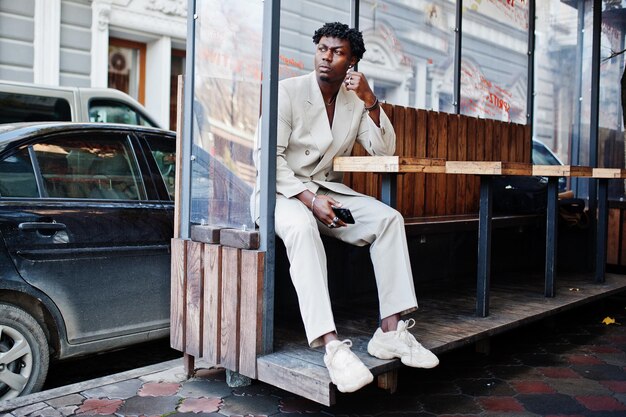 Elegante uomo afro in abito beige vecchia scuola seduto a tavola per strada Giovane maschio africano alla moda in giacca casual sul torso nudo