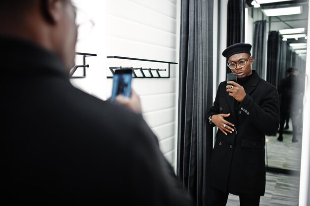 Elegante uomo afroamericano casual con berretto nero e soprabito al negozio di vestiti per camerini che fa foto allo specchio sulla fotocamera del suo telefono